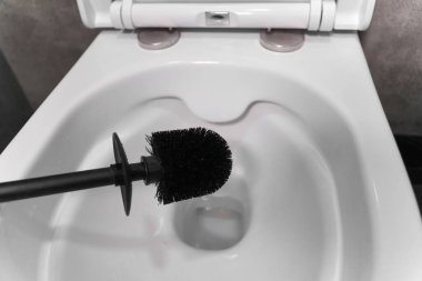 Siyah tuvalet fırçası açık, el değmemiş beyaz bir klozetin üzerinde süzülüyor, banyo hijyeninin önemini vurguluyor ve temizlik ve tazelik duygusunu teşvik ediyor.