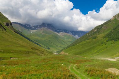 Manzarada, yuvarlanan tepeler arasında oyulmuş canlı yeşil bir vadi beyaz bulutlarla dolu dinamik bir gökyüzü altında görkemli kar örtülü zirvelere doğru yol alıyor.