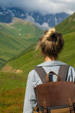 Sırt çantalı kadın yürüyüşçü, Gürcistan 'ın svaneti bölgesindeki kar kaplı dağların ve yeşil vadilerin çarpıcı manzarasının keyfini çıkararak doğanın güzelliğini ve macera ruhunu gözler önüne seriyor.