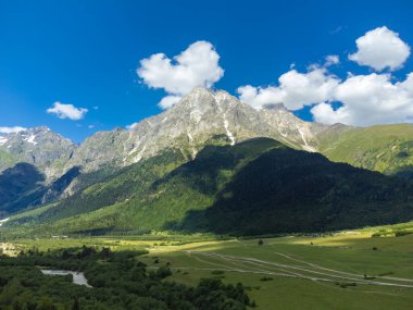 Görkemli bir dağ zirvesinin nefes kesici panoramik manzarası canlı yeşil bir vadi üzerine gölgeler döküyor ağaçlar ve parlak mavi bir gökyüzünün altında dönen bir nehir.