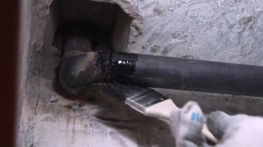 İnşaat işçisi koruyucu eldivenler giyip korozyonu önlemek ve yaşam sürelerini uzatmak için metal borulara siyah zift boyayı dikkatlice uyguluyor.