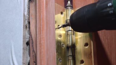 Marangoz ellerine yakın çekim, ahşap kapıya altın menteşe takmak için kablosuz matkabı ustalıkla kullanma, vida veya kapı vidaları.