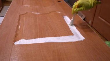 Deneyimli koruyucu sarı eldivenler giyen deneyimli bir ressam ahşap kapı paneline beyaz boya sürerek yenileme evindeki karmaşık yüzey detaylarını ortaya çıkarıyor. Kahverengi kapıları beyaza boyuyor.