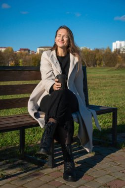 İş kadını, huzurlu bir anın tadını çıkarırken, sonbahar yeşilliklerinin ılık renkleriyle çevrili bir bankta, sakin kentsel manzarayı seyrederken bir yandan da bir parkın tepesinde kahvesini yudumluyor. Dikey fotoğraf