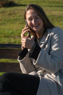 Genç, gülümseyen bir kadın, parktaki bankta oturup güneşli bir sonbahar gününün tadını çıkarırken akıllı telefonuyla mutlu bir şekilde konuşuyor. Dikey fotoğraf