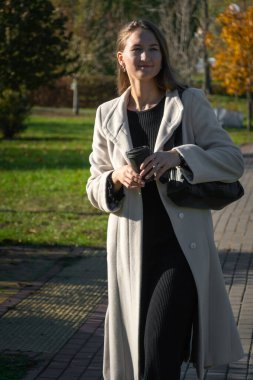 Açık renk ceketli genç bir kadın elinde bir fincan kahveyle parkta geziniyor ve güzel bir sonbahar gününde molasının tadını çıkarıyor. Dikey fotoğraf