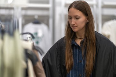 Kıyafet seçen, moda mağazasında alışveriş yapan, elbise rafının yanında duran, giysileri inceleyen, satın alma kararı veren şık bir kadın.