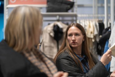 İki genç kadın bir giyim mağazasında gelecekteki alışverişleri tartışıyorlar..