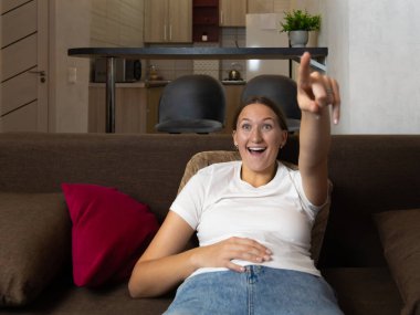 Rahat bir kanepede uzanan heyecanlı genç kadın parmağını televizyonu işaret ederek büyüleyici bir filmden ya da modern dairesinde gösteriden zevk alıyor.