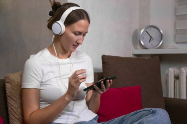 Beyaz kulaklık takan genç bir kadın oturma odasında rahat bir kanepede otururken müzik dinliyor ve akıllı telefonunu karıştırıyor.