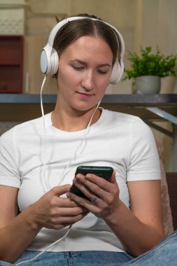 Favori çalma listesinin tadını çıkaran, kulaklık takan genç bir kadın evinde akıllı telefonunu karıştırıyor, rahatlatıcı bir deneyim için müzik ve mobil teknolojiyi birleştiriyor.