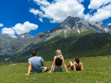 Üç turist çimenli bir çayırda soluk soluğa kalmış, parlak mavi bulutlu svaneti dağının altında Tetnuldi Dağı 'nın görkemli manzarasında dinleniyor.