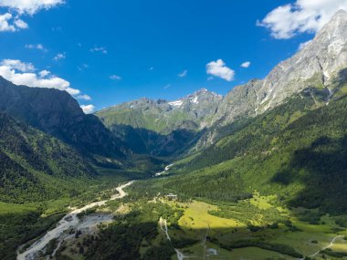Parlak mavi gökyüzü altında tüylü beyaz bulutlar oluşturan, yemyeşil ormanların arasından akan bir nehrin olduğu pitoresk bir dağ vadisinin havadan görünüşü.