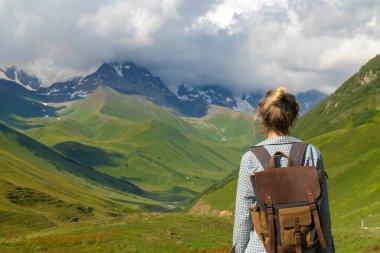 Sırt çantalı kadın yürüyüşçü, yeşil vadi ve karlı tepelerin nefes kesici manzarasının keyfini çıkarıyor. Dağ macerası sırasında doğanın güzelliğini kucaklıyor.