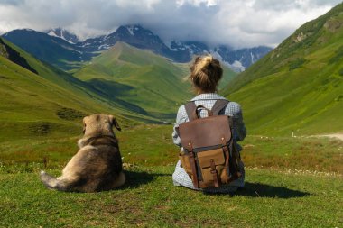Sırt çantalı bayan yürüyüşçü köpeğiyle çimenli dağ yamacında dinleniyor, yaz macerası boyunca karlı tepelerin ve yeşil vadilerin nefes kesici manzarasının keyfini çıkarıyor.