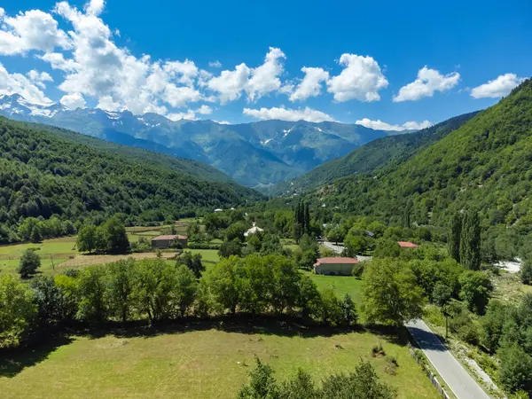 Yeşil çayırlar, yemyeşil ormanlar, evler, dolambaçlı bir yol ve beyaz bulutlu parlak mavi bir gökyüzünün altında kar kaplı zirveler olan pitoresk bir dağ vadisi.