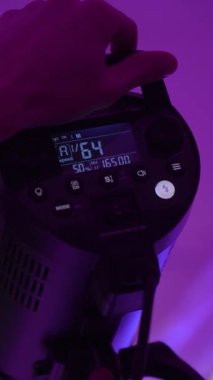 Profesyonel fotoğrafçı, loş mor ışıklı stüdyoda kamera flaş ayarlarını ayarlıyor titizlikle profesyonel fotoshoot 'u ileri düzey ışıklandırma ekipmanıyla dikey video ile hazırlıyor.