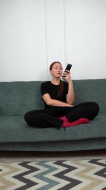 Rahat bir koltukta oturan, günlük kıyafetler giyen uzaktan kumanda kullanan, televizyon izleyen ilginç bir şey arayan genç bir kadın. Dikey video