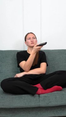 Rahat bir koltukta oturan, günlük kıyafetler giyen uzaktan kumanda kullanan, televizyon izleyen ilginç bir şey arayan genç bir kadın. Dikey video