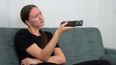 Genç bir kadın rahat bir koltukta oturuyor, arızalı bir TV kumandasını çalıştırmaya çalışırken hayal kırıklığı yaşıyor, düğmelere basıyor ve kanal değiştiriyor.