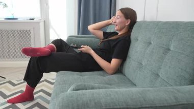 Genç kadın rahat bir şekilde kanepede oturuyor, televizyon izliyor uzaktan kumandayla kanal değiştiriyor, gülüyor ve evde boş zamanlarının tadını çıkarıyor.