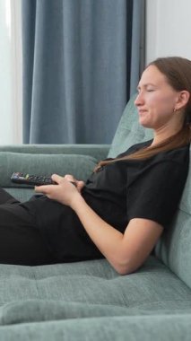 Genç kadın kanepede rahat bir şekilde oturdu, televizyon izledi, uzaktan kumandayla kanal değiştirdi, güldü ve evde boş zamanlarının tadını çıkardı. dikey video.