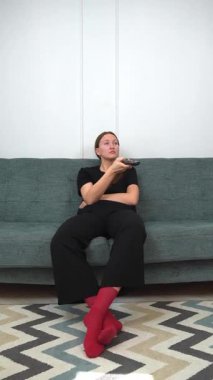 Rahat bir koltukta oturan, günlük kıyafetler giyen uzaktan kumanda kullanan, televizyon izleyen ilginç bir şey arayan genç bir kadın. Dikey video