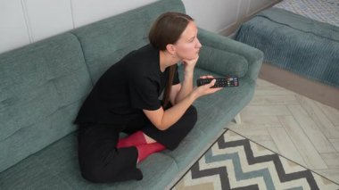 Genç bir kadın rahat bir şekilde kanepede oturuyor, uzaktan kumandayla televizyon kanallarında geziniyor, oturma odasındaki içeriği arıyor.