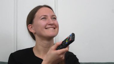 Kanepede oturan, elinde uzaktan kumandayla televizyon izleyen, kanal değiştiren, gülümseyen, beyaz bir arka planda izole edilmiş genç bir kadın.