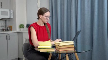 Evde öğrenim ortamında kırmızı kolsuz bluz ve gözlük takan, etrafı kitaplarla, laptoplarla çevrili cam masada çalışırken yeşil kalemle notlar alan odaklanmış genç bir öğrenci.