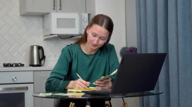 Kişisel mali durumları analiz eden, bütçeyi yöneten, nakit sayan, modern mutfak ortamında çalışırken dizüstü bilgisayarına detaylı notlar alan genç profesyonel bir kadın.