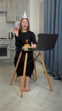 Renkli parti şapkalı genç bir kadın doğum gününü neredeyse kutluyor, pasta yerken korna çalıyor ve dizüstü bilgisayardan ev mutfağından video arıyor. dikey video