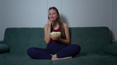 Genç bir kadın film gecesinden zevk alıyor, yeşil kanepede oturuyor ve patlamış mısırın tadını çıkarıyor.
