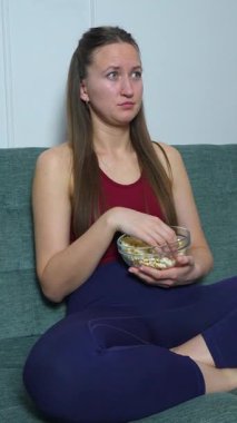Kanepede oturup film izleyen ve cam kaseden patlamış mısır yiyen genç bir kadın mutluluktan üzüntüye, dikey videoya çeşitli duygular gösteriyor.