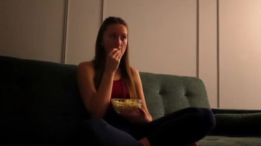 Kanepede oturan genç bir kadın, film izliyor ve cam kaseden patlamış mısır yiyor, mutluluktan üzüntüye kadar çeşitli duygular gösteriyor.
