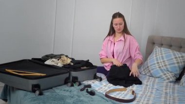 Giysileri ve seyahat aksesuarlarını dikkatlice düzenleyen genç bir kadın yaklaşan yolculuk için bavulunu hazırlarken, evden çıkmadan önce rahat bir şekilde yatağında oturuyor.