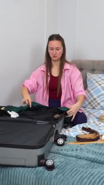 Genç kadın giysilerini ve seyahat aksesuarlarını dikkatlice düzenliyor, giysilerini düzgünce katlıyor ve yaklaşan dikey yolculuk videosu için hazırlanırken eşyaları bir bavula koyuyor.