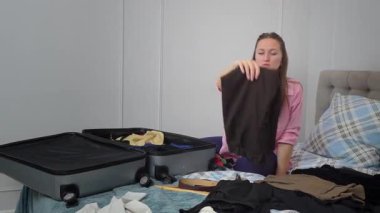 Neşeli bayan turist yaz tatili kıyafetlerini açık bavula yerleştiriyor, rahat bir şekilde yatakta oturuyor ve yaklaşan yolculuk için kişisel eşyaları düzenliyor.