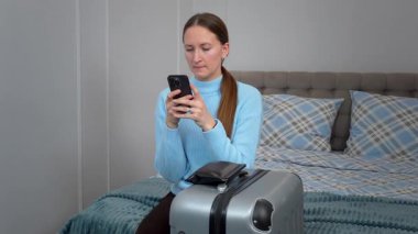 İş seyahatine ya da tatile hazırlanan genç bir kadın bavullarıyla yatakta oturuyor, akıllı telefonu seyahat planlama ve internet iletişimi için kullanıyor ya da taksi çağırıyor.