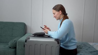 İş seyahatine ya da tatile hazırlanan genç bir kadın bavullarıyla yatakta oturuyor, akıllı telefonu seyahat planlama ve internet iletişimi için kullanıyor ya da taksi çağırıyor.