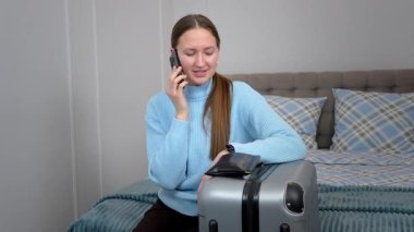 Genç Bayan Gezgin, Bavulun yanındaki otelde oturuyor, cep telefonuyla konuşuyor, seyahatin detaylarını planlıyor ve yaklaşan yolculuk için ulaşımı organize ediyor.