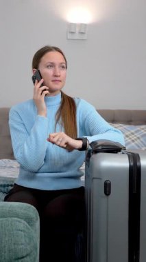 Endişeli turist kadın bavuluyla bir otel odasında oturuyor, telefonla konuşuyor ve akıllı saatini kontrol ediyor, taksi ya da mekiğin dikey videosunu bekliyor.