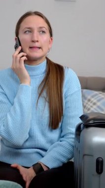 Endişeli turist kadın bavuluyla bir otel odasında oturuyor, telefonla konuşuyor ve akıllı saatini kontrol ediyor, taksi ya da mekiğin dikey videosunu bekliyor.