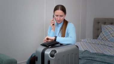 Endişeli turist kadın bavuluyla bir otel odasında oturuyor, telefonla konuşuyor ve akıllı saatini kontrol ediyor, taksisini ya da mekiğini bekliyor.
