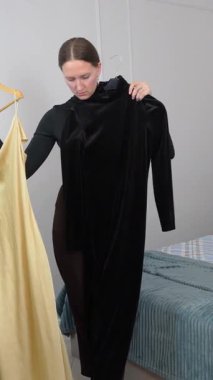 Göz kamaştırıcı kadın yatak odasında dikiliyor, altın saten iç çamaşırı ve askılarda siyah kadife elbiseyi dikkatlice karşılaştırıyor, moda karar verme anını dikey videoda yaşıyor.