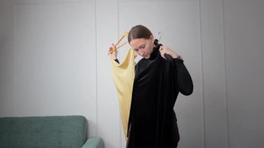 Genç bir kadın yatak odasında dikiliyor, sarı elbise ve siyah tulumu askılarda asılı halde dikkatlice karşılaştırıyor, gelecek etkinlik için kıyafet seçmeden önce moda seçimini düşünüyor.
