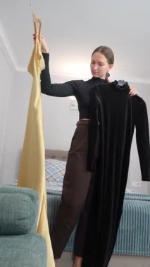 Yatak odasında duran genç bir kadın, sarı elbise ve siyah tulumu askılarda asılı halde dikkatlice karşılaştırıyor, yaklaşan olay videosu için kıyafet seçmeden önce moda seçimini düşünüyor.