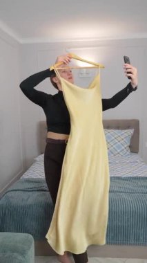 Moda blogcusu, yatak odasında akıllı telefon özçekimleri çekerken askıda uzun sarı elbiseler sergiliyor, çevrimiçi kıyafet satışları için popüler sosyal medya içeriğini dijital video satışları için yakalıyor