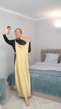 Moda blogcusu, yatak odasında akıllı telefon özçekimleri çekerken askıda uzun sarı elbiseler sergiliyor, çevrimiçi kıyafet satışları için popüler sosyal medya içeriğini yakalıyor
