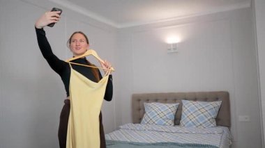 Moda blogcusu, yatak odasında akıllı telefon özçekimleri çekerken askıda uzun sarı elbiseler sergiliyor, çevrimiçi kıyafet satışları için popüler sosyal medya içeriğini yakalıyor